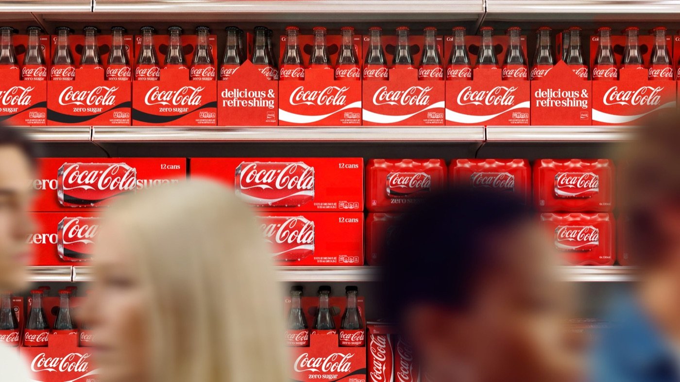 Rayon de supermarché rempli de packs Coca-Cola rouges, avec le ruban blanc en grand format et la mention delicious and refreshing en serif